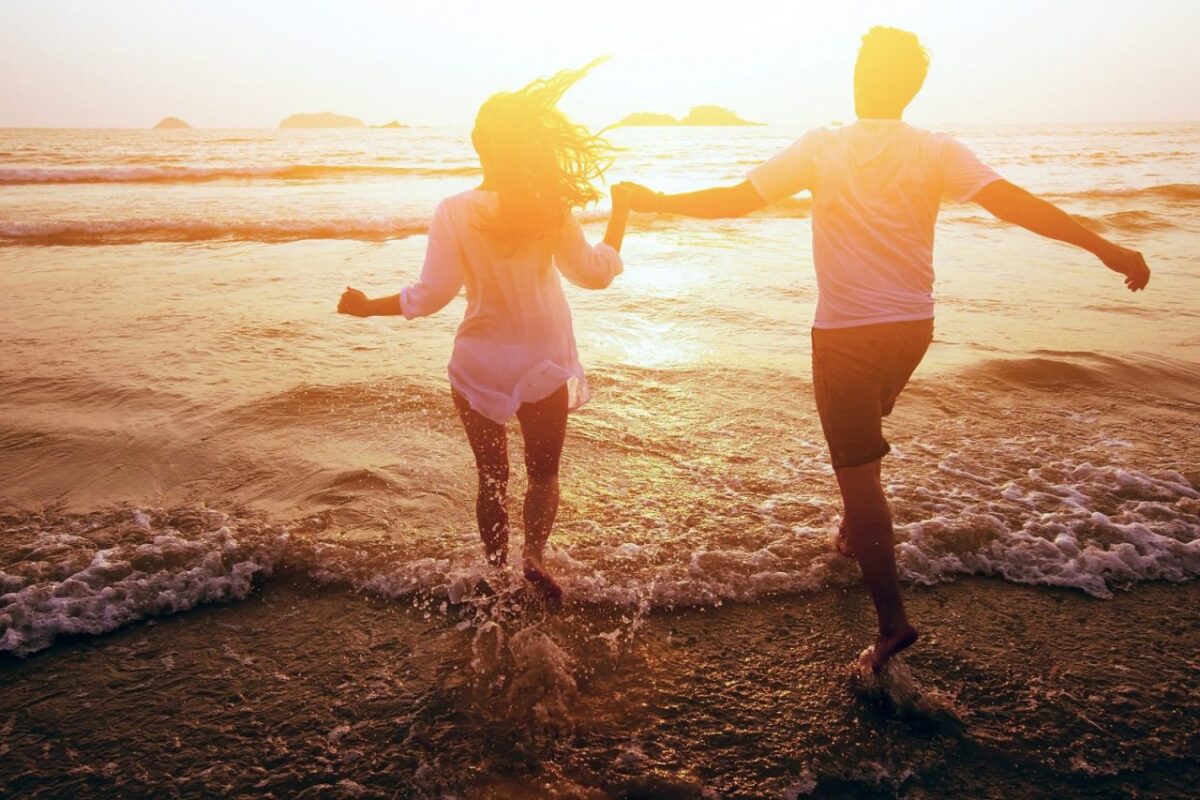 A man and a woman holding hands and running through shallow ocean water at sunset, with islands visible in the distance.