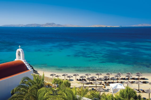 A scenic view of a beach with clear turquoise waters, lined with umbrellas and sun-loungers, and a church with a red roof in the foreground.