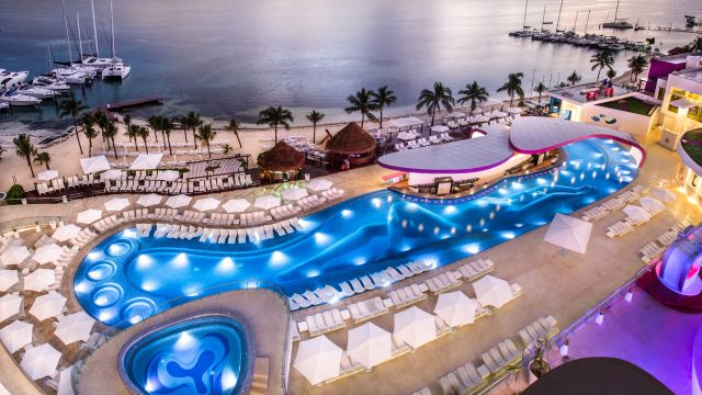 Aerial view of a lit-up, serpentine pool beside a beach with umbrellas at dusk, overlooking a tranquil marina with yachts.