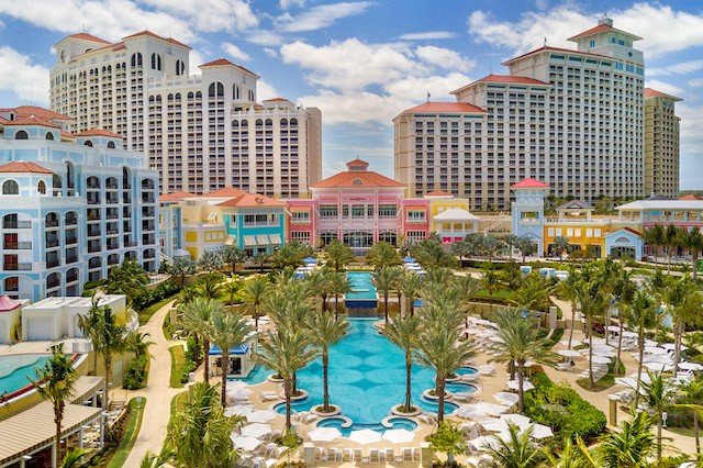 Aerial view of a luxury resort with multiple high-rise buildings and a central swimming pool flanked by palm trees.