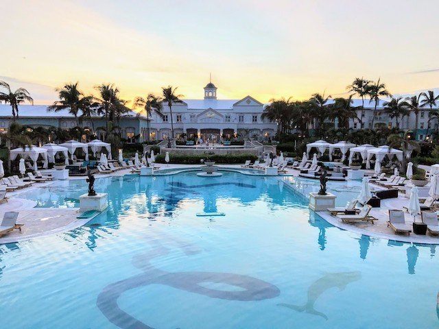Luxury resort with a large swimming pool at dusk, surrounded by white loungers and a grand white building with a central dome in the background.