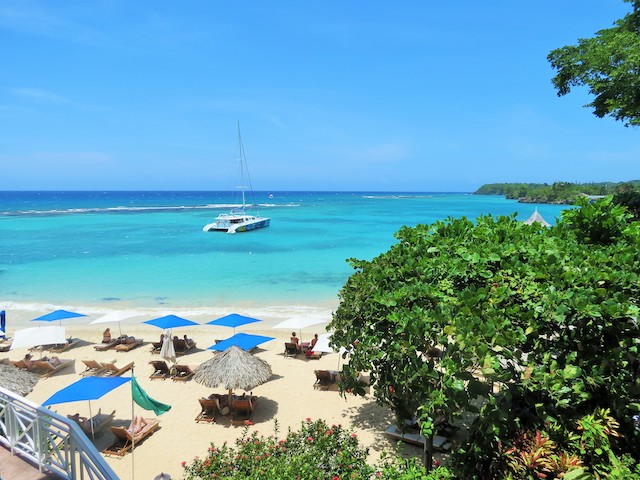 A beach scene with sun loungers, umbrellas, and a sailboat on turquoise waters under a clear blue sky. Trees and bushes occupy part of the foreground.