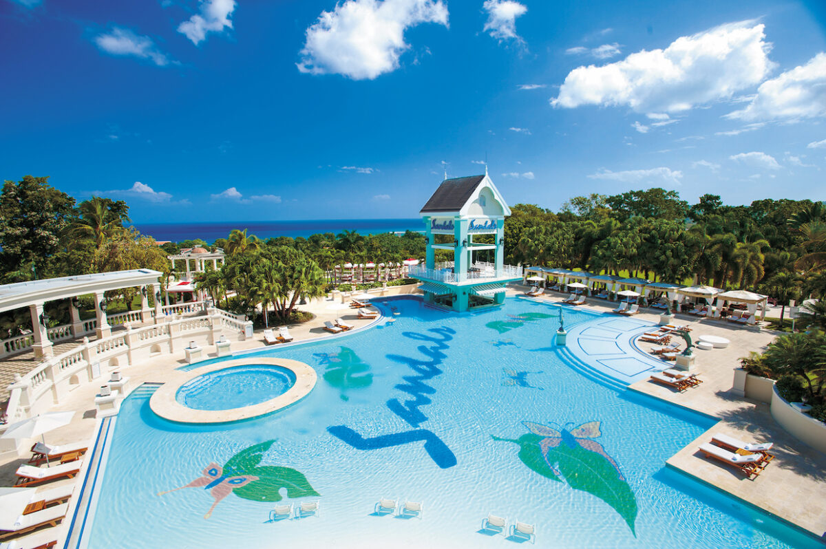 A large resort swimming pool with "Sandals" written on the bottom, surrounded by lounge chairs, cabanas, and tropical vegetation, with the ocean visible in the background under a clear blue sky.