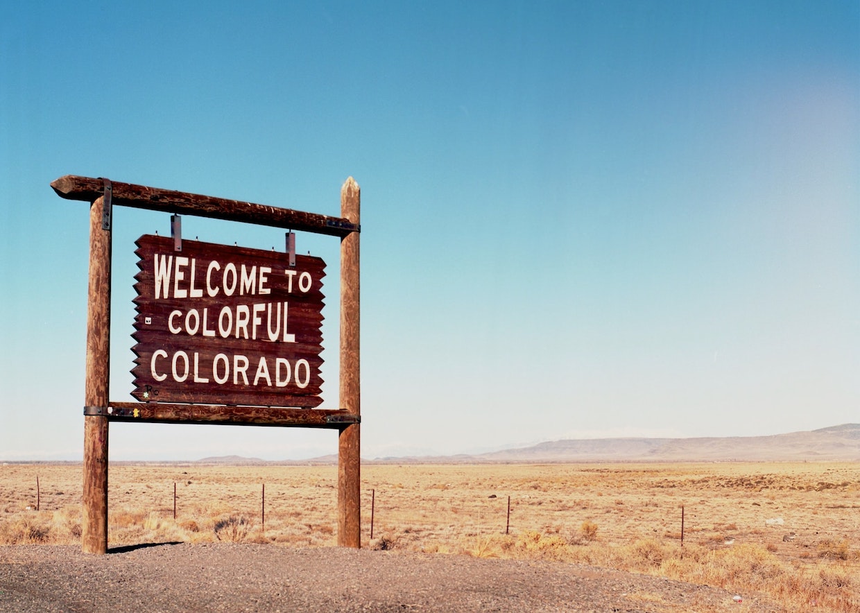 Wooden roadside sign reading "Welcome to Colorful Colorado" in a desert landscape under a clear blue sky.