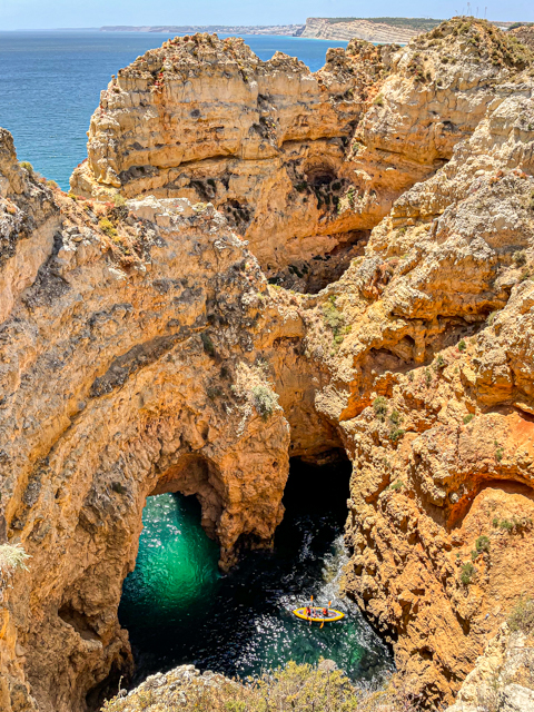 A yellow kayak navigates through a natural rock archway in a coastal cliff formation with blue-green water below and clear skies above.
