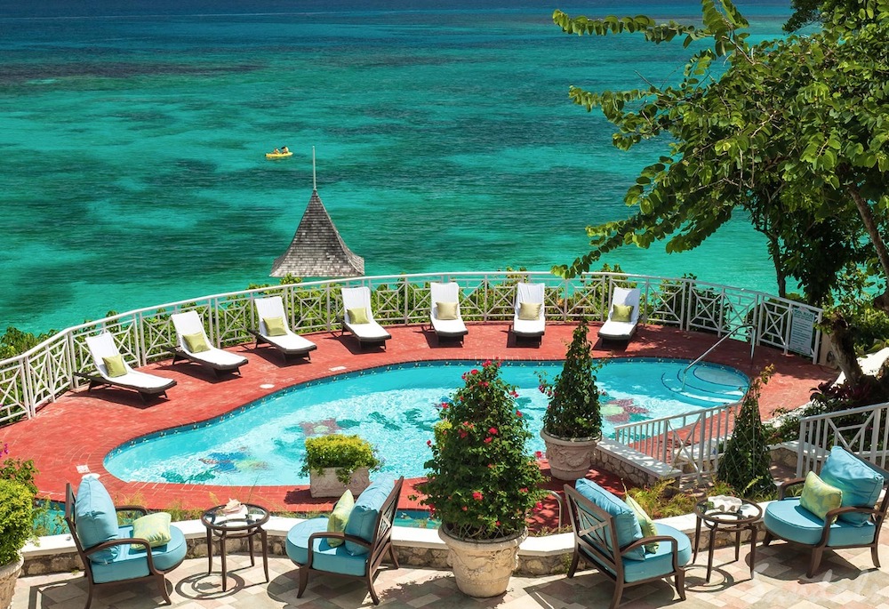 A pool terrace with lounge chairs overlooks a clear, turquoise ocean. Trees and lush greenery surround the area. A small gazebo-like structure is visible near the shore.