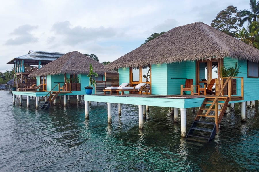 Two turquoise overwater bungalows with thatched roofs stand on stilts above clear water, each with a deck, chairs, and access steps. Lush trees are in the background.