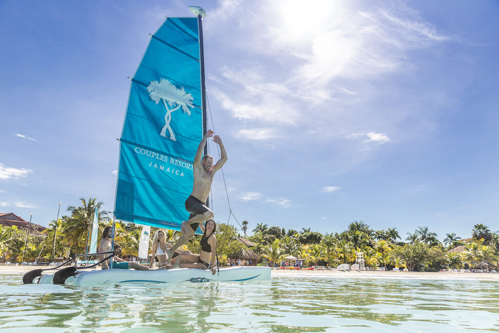 A man jumps from a sail boat into the ocean. A small group of people stay on the boat and watch.