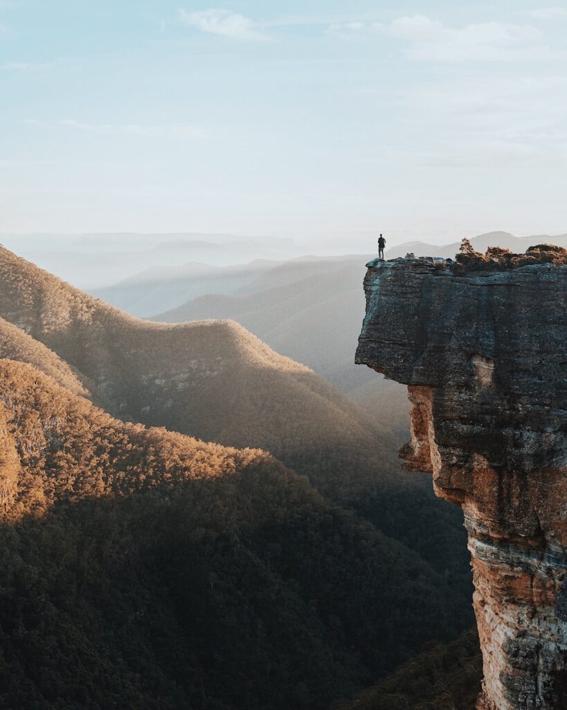 A man standing on a large cliff overlooking other mountains.