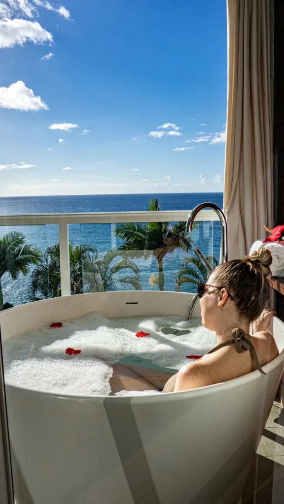 A person relaxes in a bubble bath with rose petals, overlooking an ocean view and palm trees through a glass balcony.