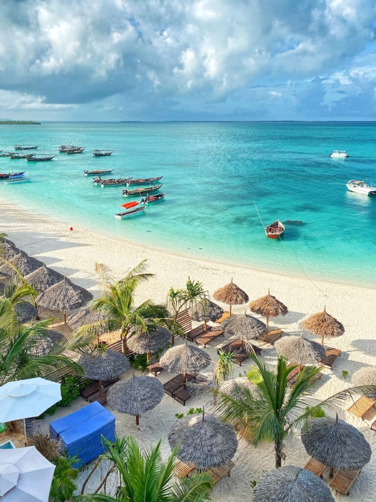 Zanzibar beach with boats in the water and thached umbrellas over lounge chairs.