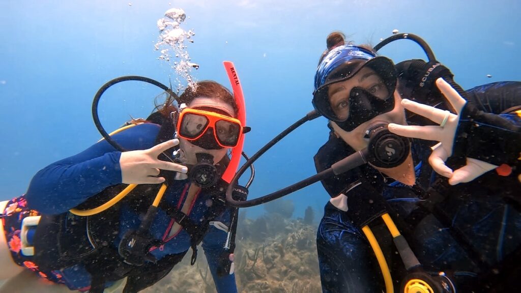 Two scuba divers underwater wearing colorful diving gear, including masks and snorkels. Both are making hand gestures while surrounded by clear blue water and coral formations.