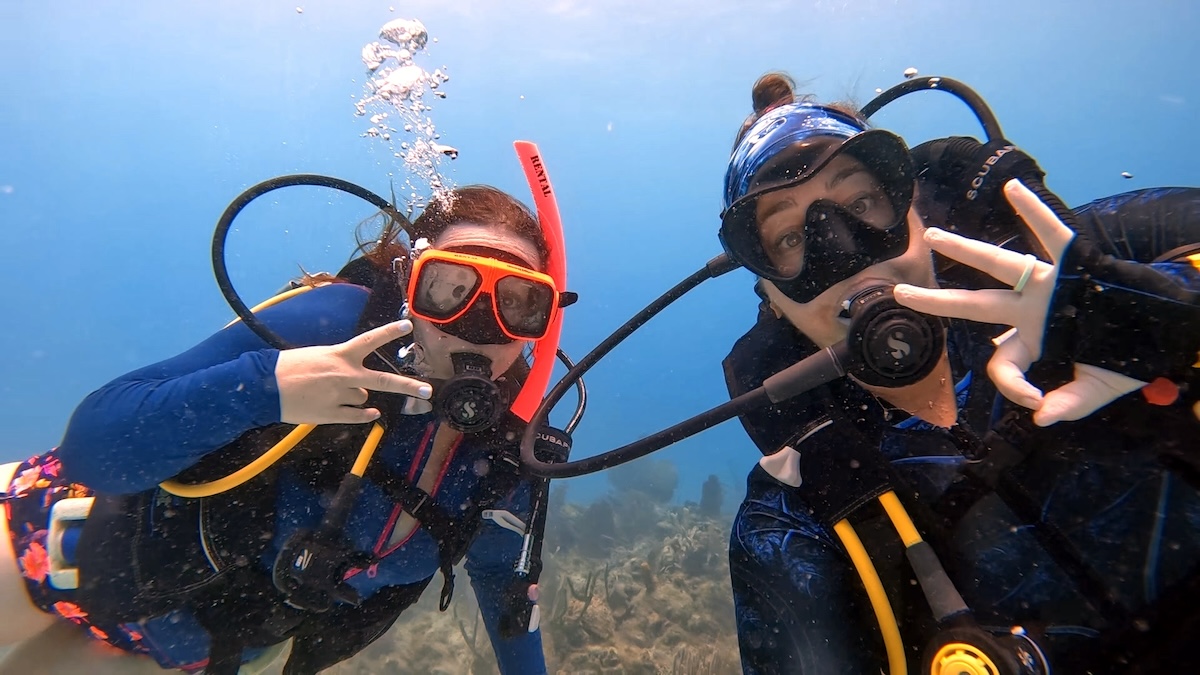 Two scuba divers underwater wearing colorful diving gear, including masks and snorkels. Both are making hand gestures while surrounded by clear blue water and coral formations.