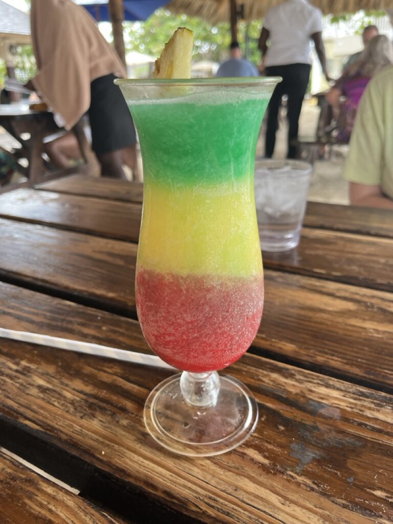 A layered cocktail in a tall glass, featuring red, yellow, and green layers, topped with a pineapple slice, positioned on a wooden table in an outdoor setting.