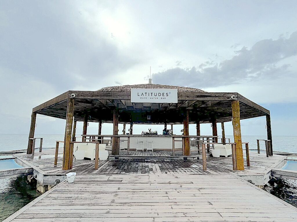 A wooden pavilion over water, labeled "Latitudes Over Water Bar", featuring a bar counter and seating area, under a cloudy sky.