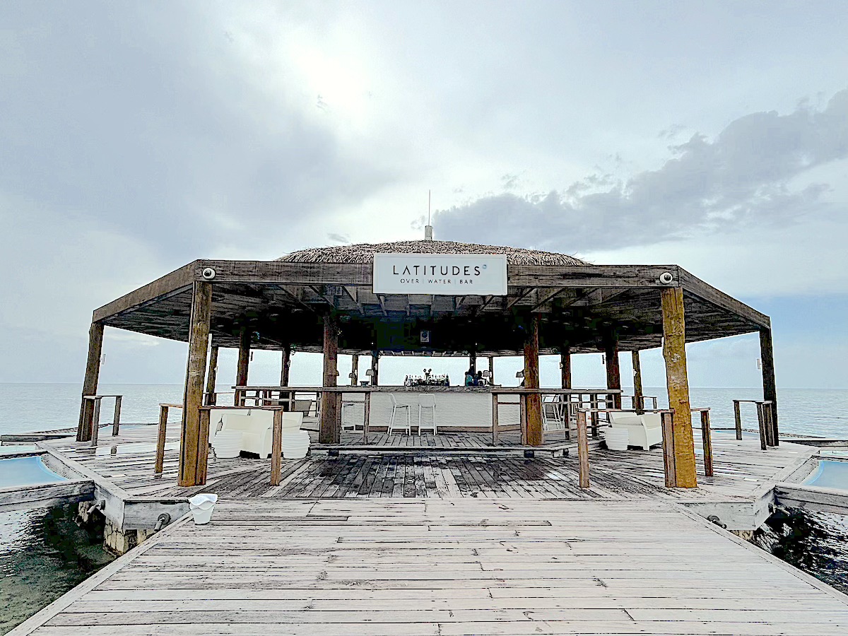 A wooden pavilion over water, labeled "Latitudes Over Water Bar", featuring a bar counter and seating area, under a cloudy sky.