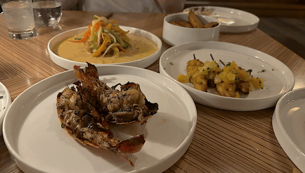 A table with several plates of food, including grilled lobster, shrimp skewers with garnish, a creamy vegetable dish, and a small bowl of breadsticks.