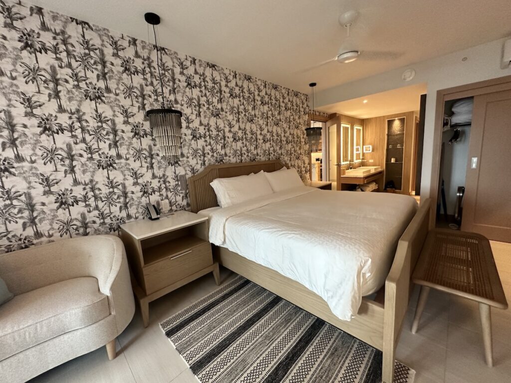 A hotel room with a double bed, a patterned accent wall, a small white armchair, a bedside table, and a bench. There is an open bathroom area visible in the background.