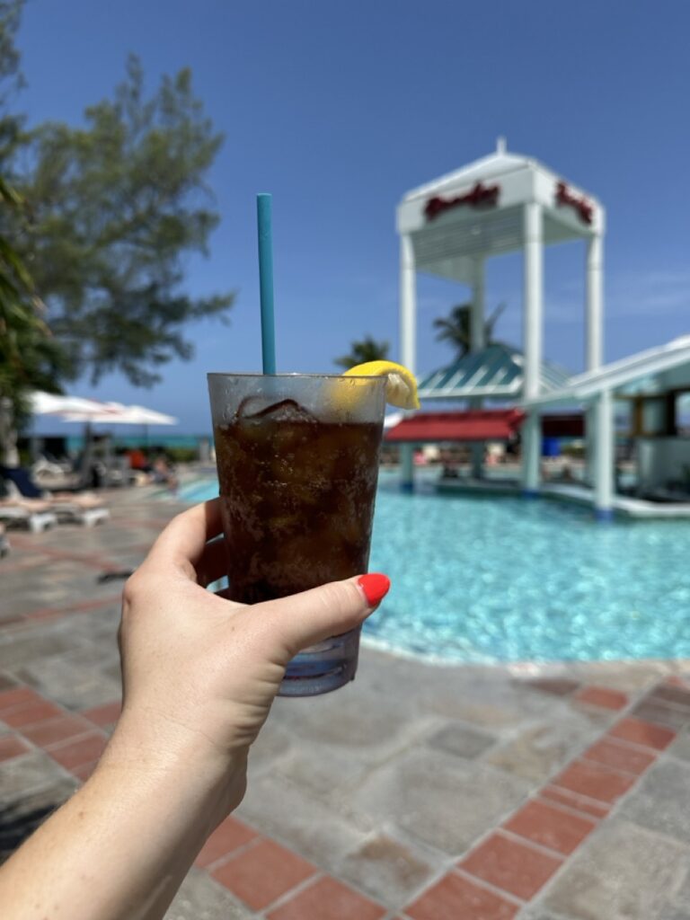 A hand holding a glass of dark soda with ice and a lemon slice near a swimming pool, with a resort structure and lounge chairs in the background.