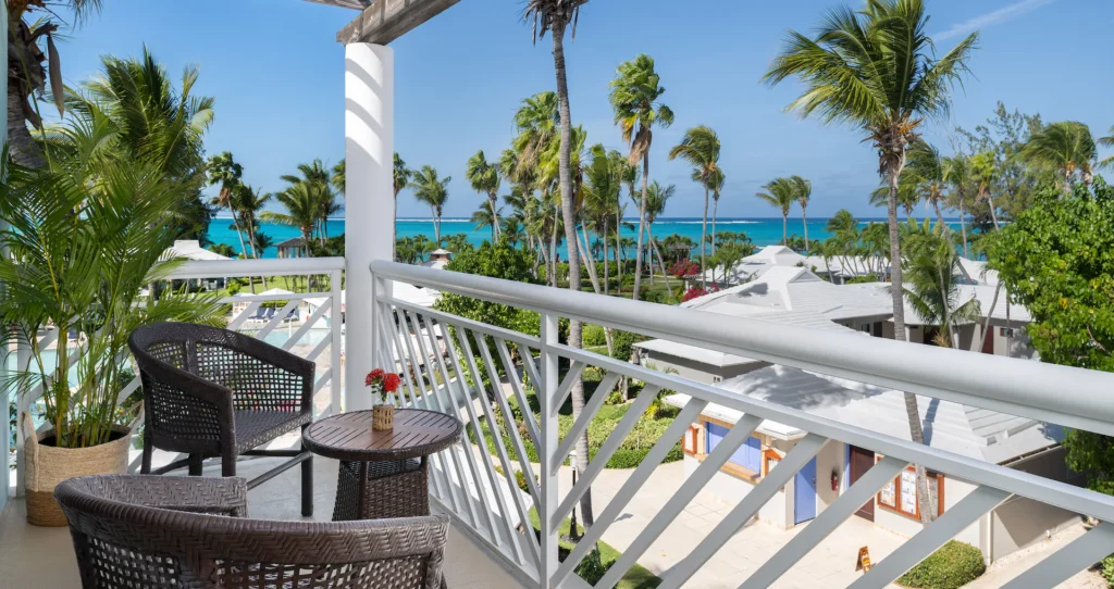 A balcony with two wicker chairs and a small table overlooks tropical palm trees, white-roofed buildings, and the blue ocean under a clear sky.