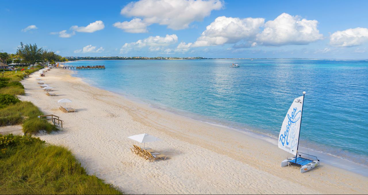 A sandy beach with lounge chairs and umbrellas along the shoreline, a sailboat onshore, calm blue water, and a partly cloudy sky.