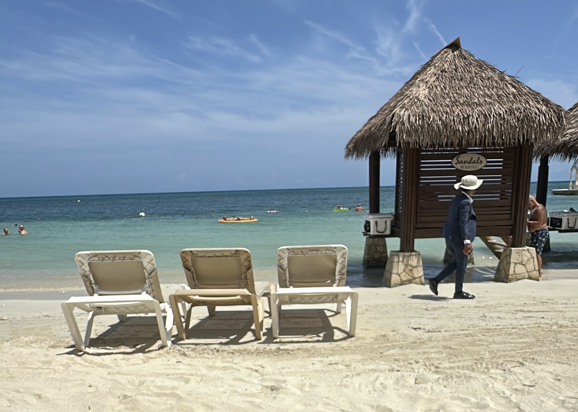 A man wearing a hat walks by four empty beach chairs and a thatched hut on a sandy beach with clear blue water in the background.
