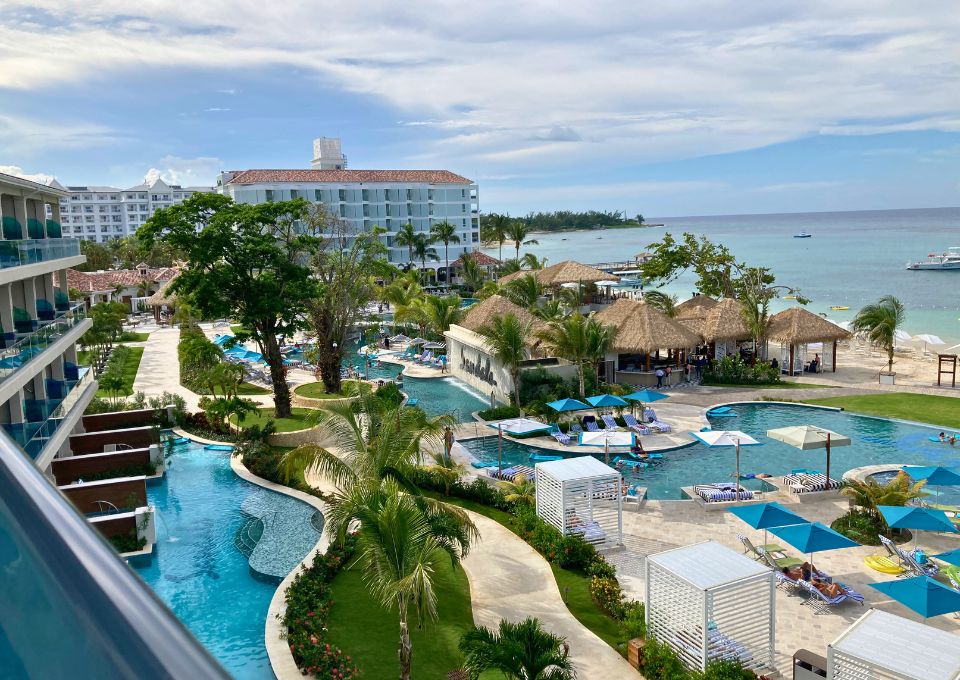 A tropical resort with a swimming pool, palm-thatched huts, and a view of the ocean.