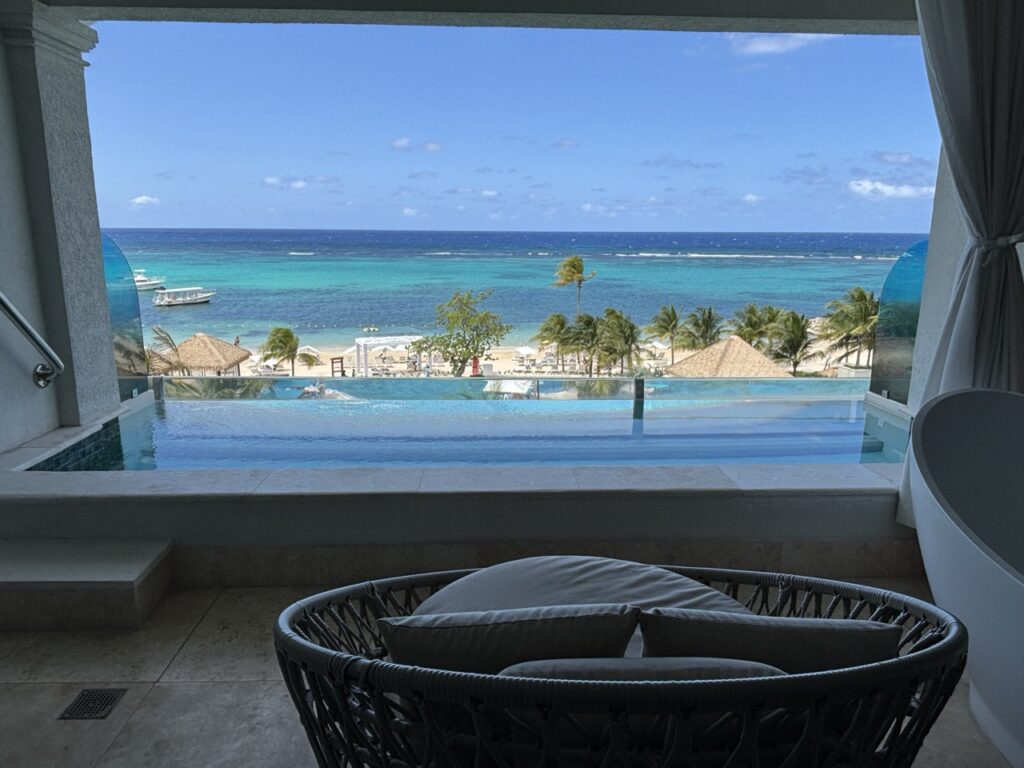 View from a shaded balcony showing a lounge chair, infinity pool, beach with palm trees, and the ocean under a blue sky.